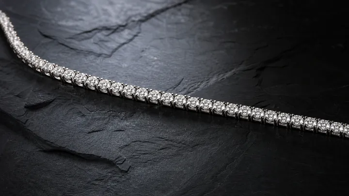 A white gold diamond tennis bracelet is draped across a dark slate surface.